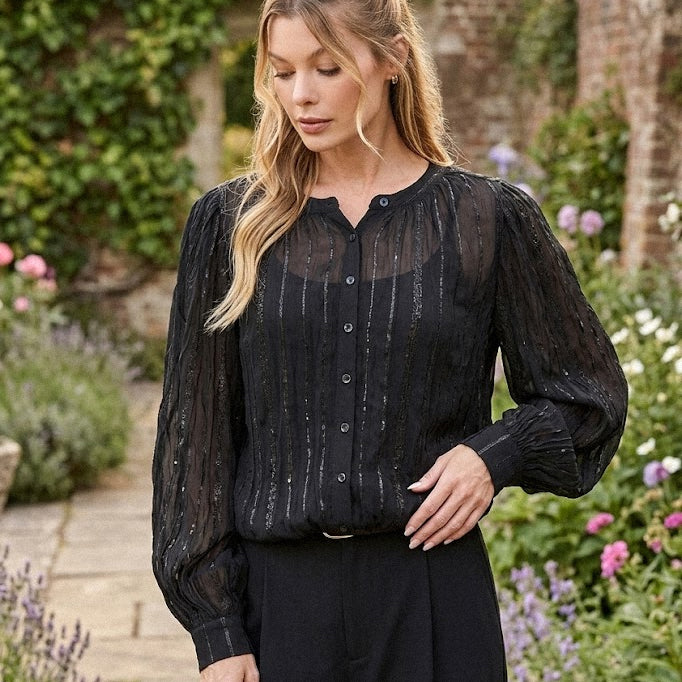A woman with long blonde hair wearing a black, slightly sheer blouse with vertical pinstripes of sequins and voluminous sleeves.