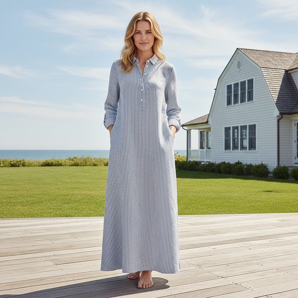 Model wearing a sophisticated navy striped European linen maxi dress with hands in pockets standing on a seaside deck.