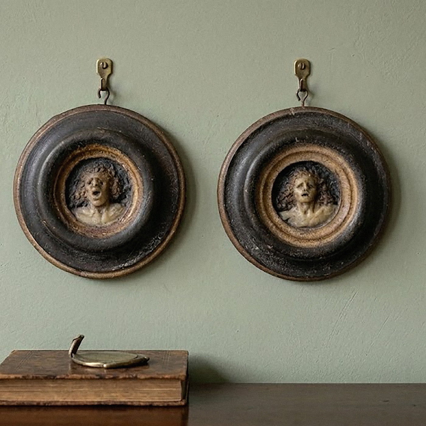 Two round wall art pieces with portraits on a green wall, with a book and decorative item at the bottom.